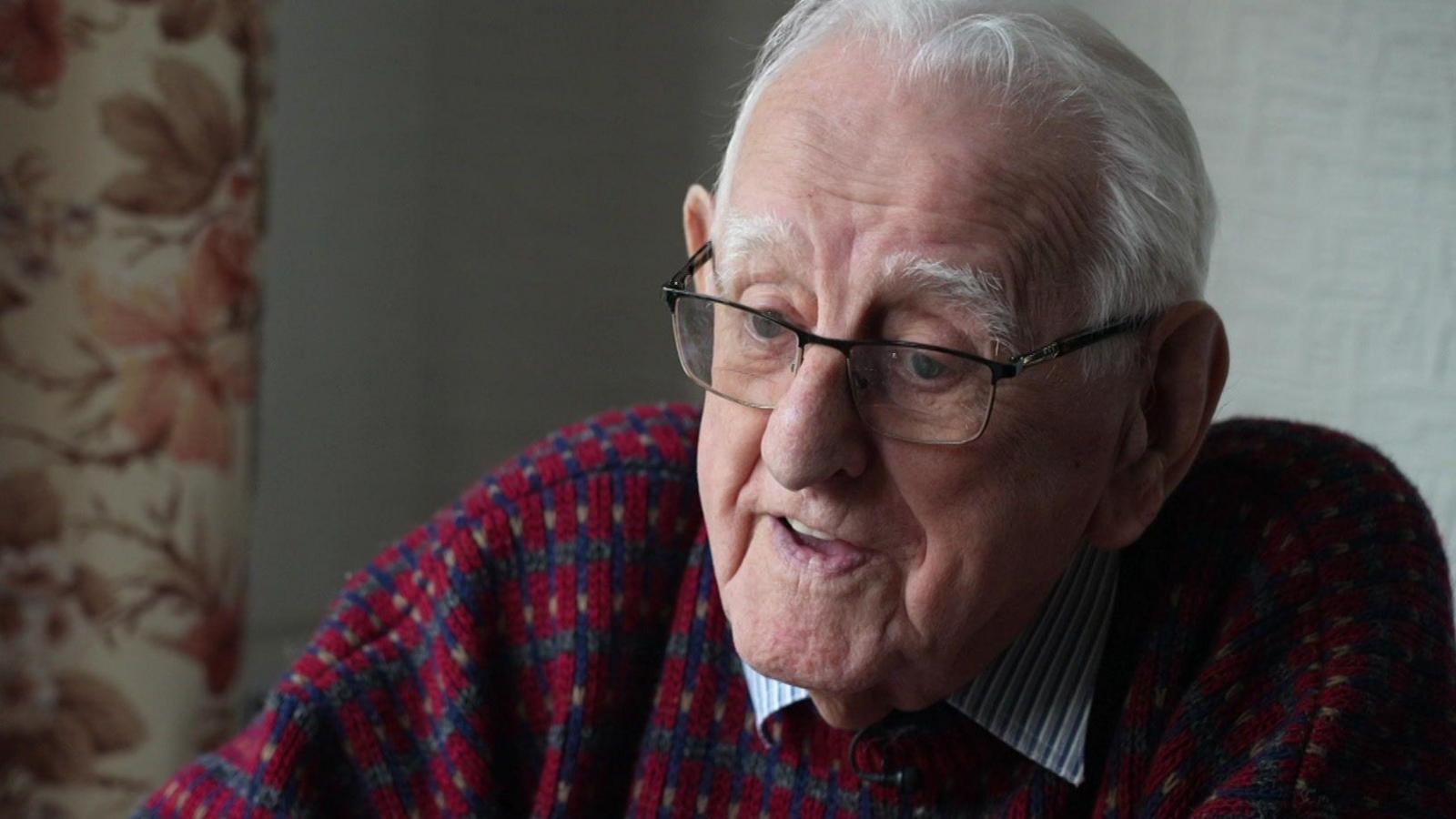 Tony Johnson wearing a blue and white shirt under a purple jumper talks at his home to the BBC