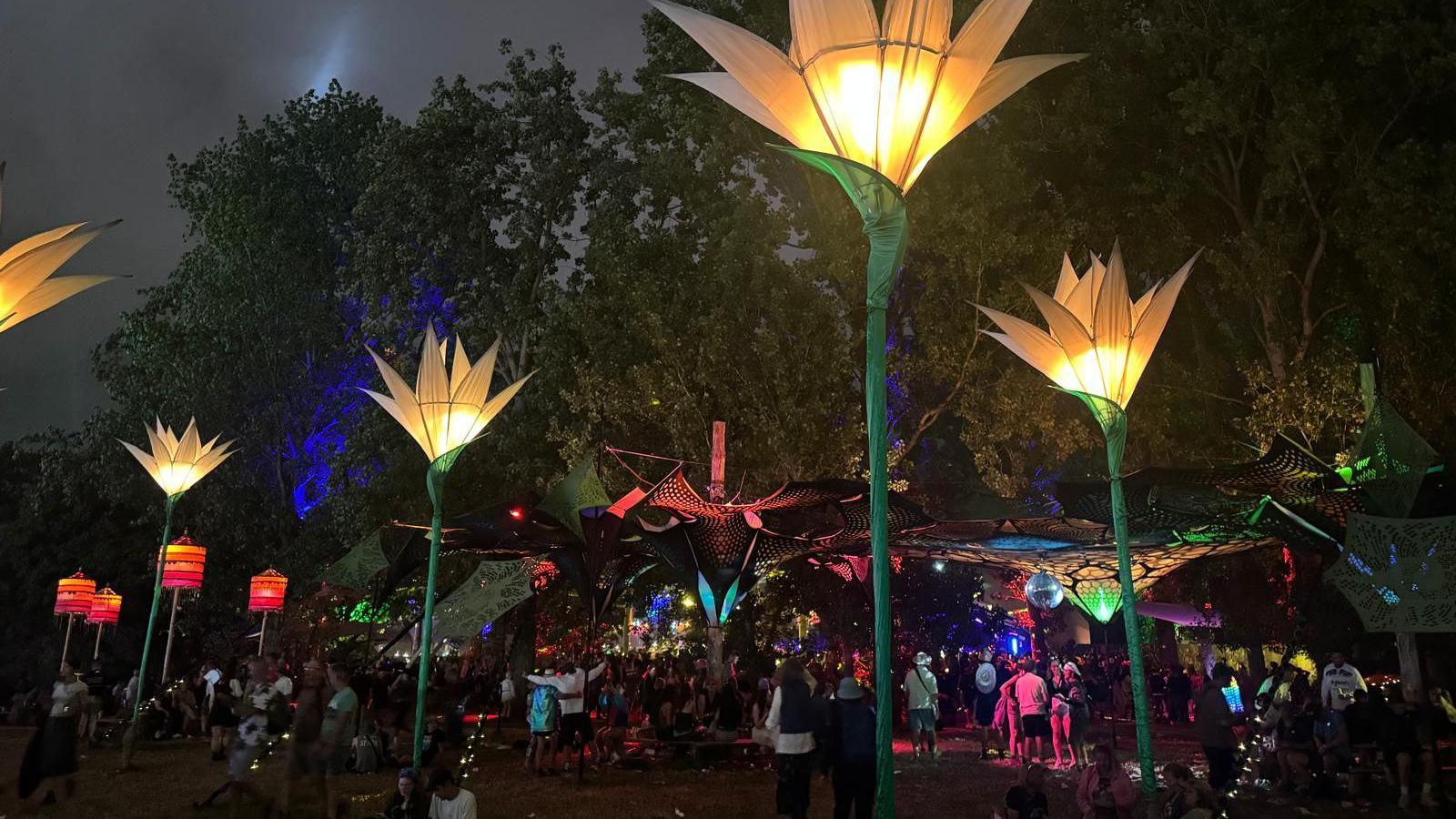 About six flower-themed lanterns can be seen dotted around an area of Glastonbury Festival. They are about the height of a street light, with green "stems" and yellow flowers. There is an under-tree canopy in the background with a disco ball hanging for it, with revellers dancing underneath it.