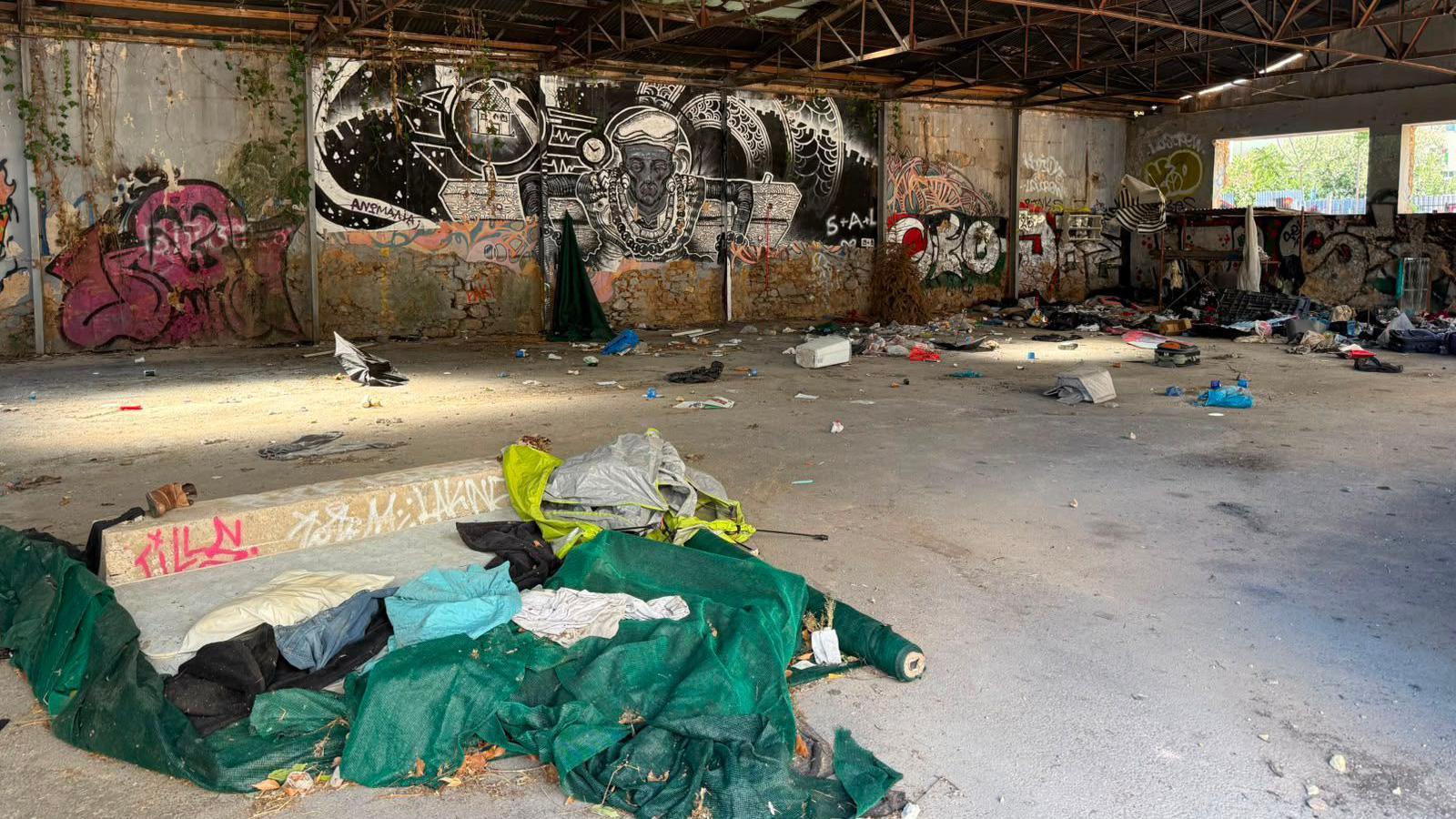 Interior of a large abandoned warehouse with graffiti-covered walls and exposed metal roof beams. The concrete floor is scattered with rubbish, clothing, and discarded materials. In the foreground, torn green tarpaulins and fabric lie beside a low concrete structure with graffiti. Large black-and-white mural artwork covers part of the back wall, while daylight enters through windows on the right side of the building.