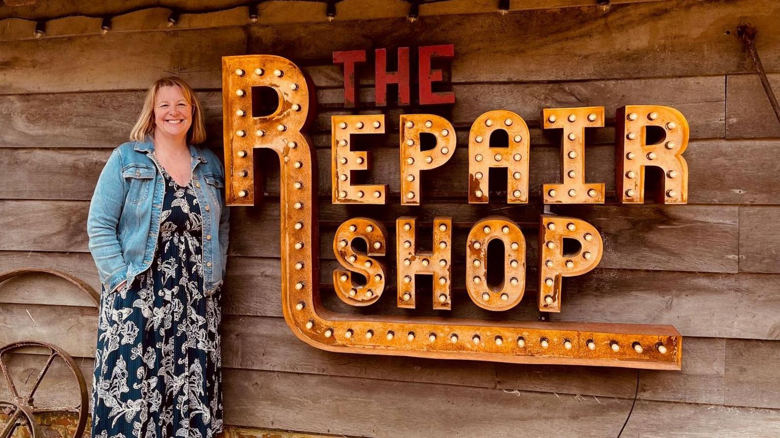 A woman wearing a blue patterened dress and a jean jacket is standing next to a sign saying The Repair Shop. The sign is made of metal and has light bulbs inside. It is hanging on a wooden shed.