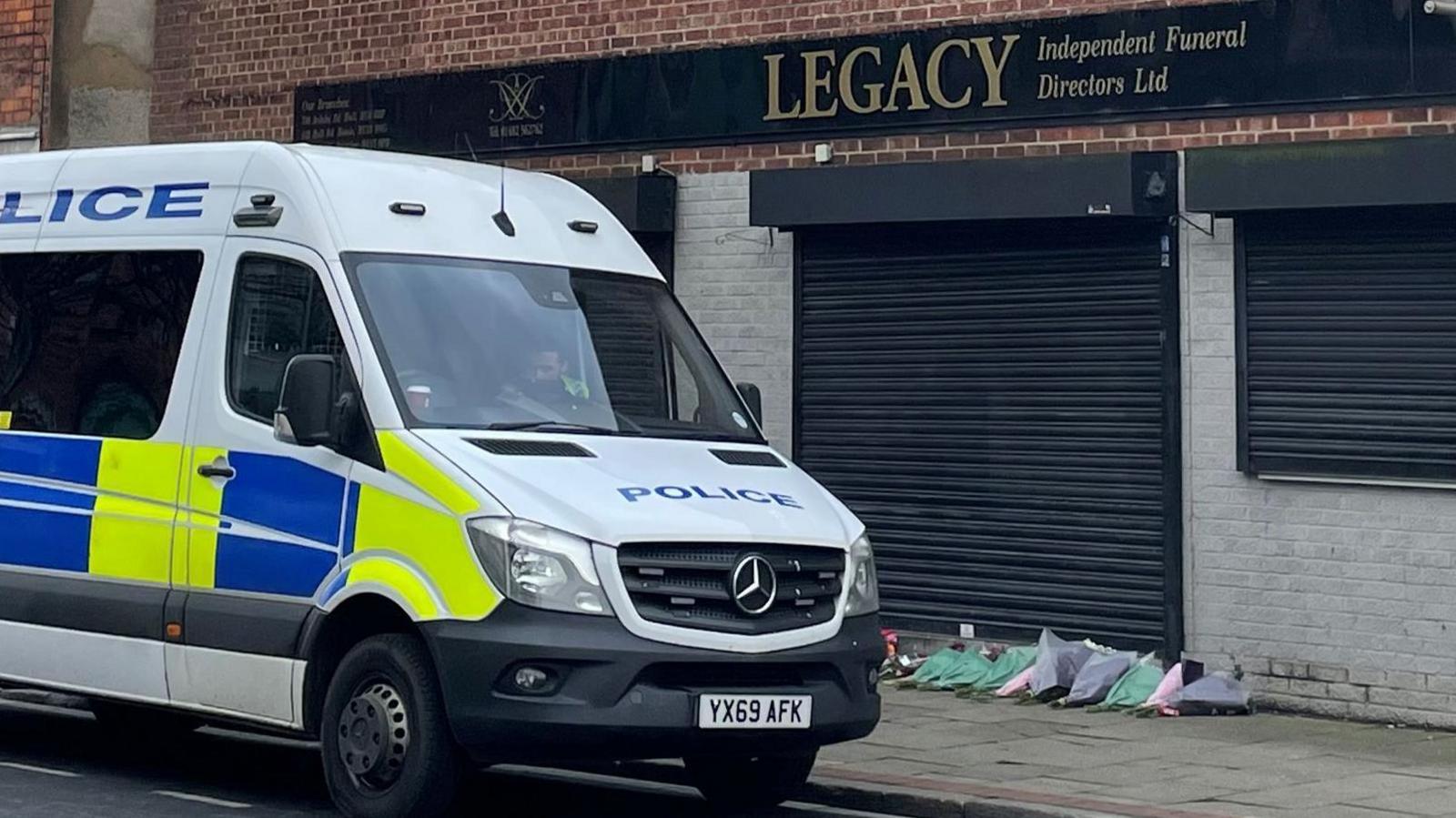 A police van parked in front of a red and grey brick building. A black sign on the front of the building reads: Legacy Independent Funeral Directors Ltd. On the ground are a dozen bouquet of lowers laid in a row against black shutters.