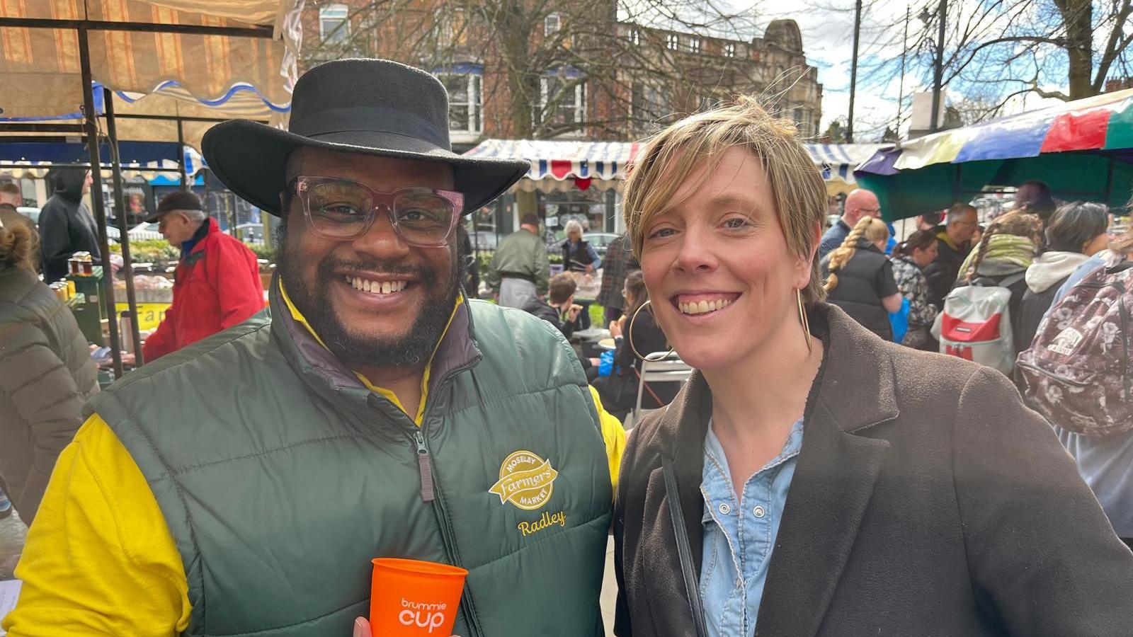 A man wearing a black hat, glasses and a green gilet is stood with a woman with short blonde hair, who is wearing a brown coat with a denim top. 
