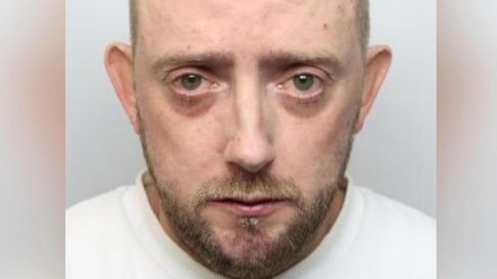 Mugshot of a mostly bald man. He looks sad and like he may have been crying, with dark circles under his eyes. He has a short light-brown beard and wears a white jumper.