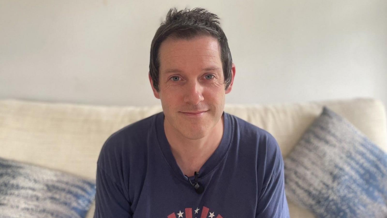 A man with short brown hair and a blue t-shirt sits on a cream sofa, looking directly into the camera. Behind him is a white wall, on the sofa there are blue cushions, one either side of him.