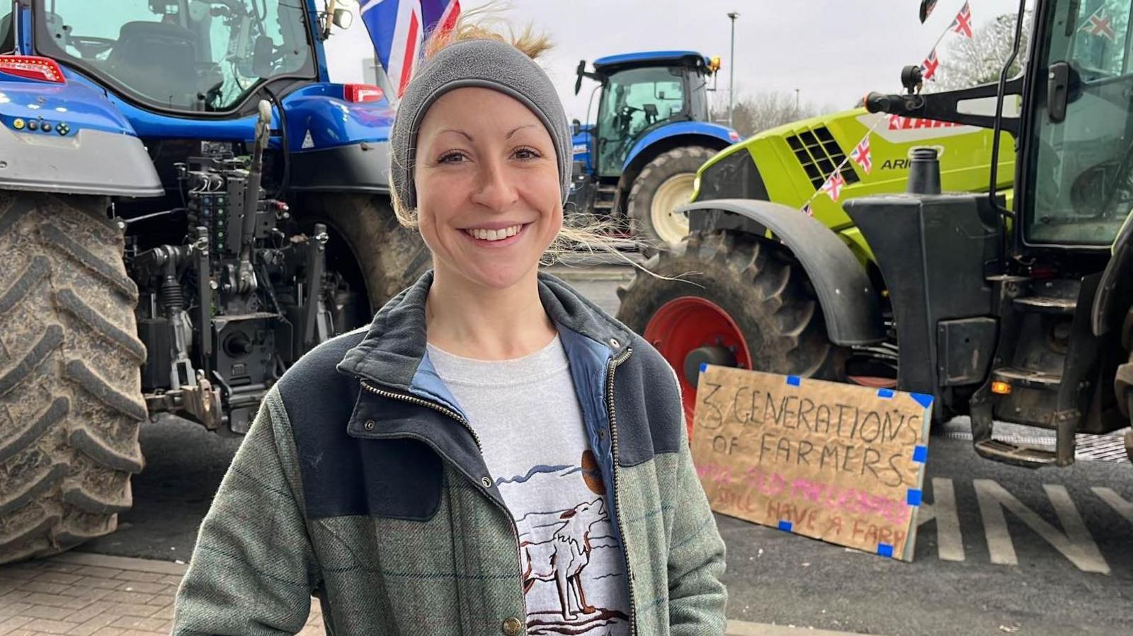 A woman in a tweed jacket, grey jersey and hat stands in front of tractors. 