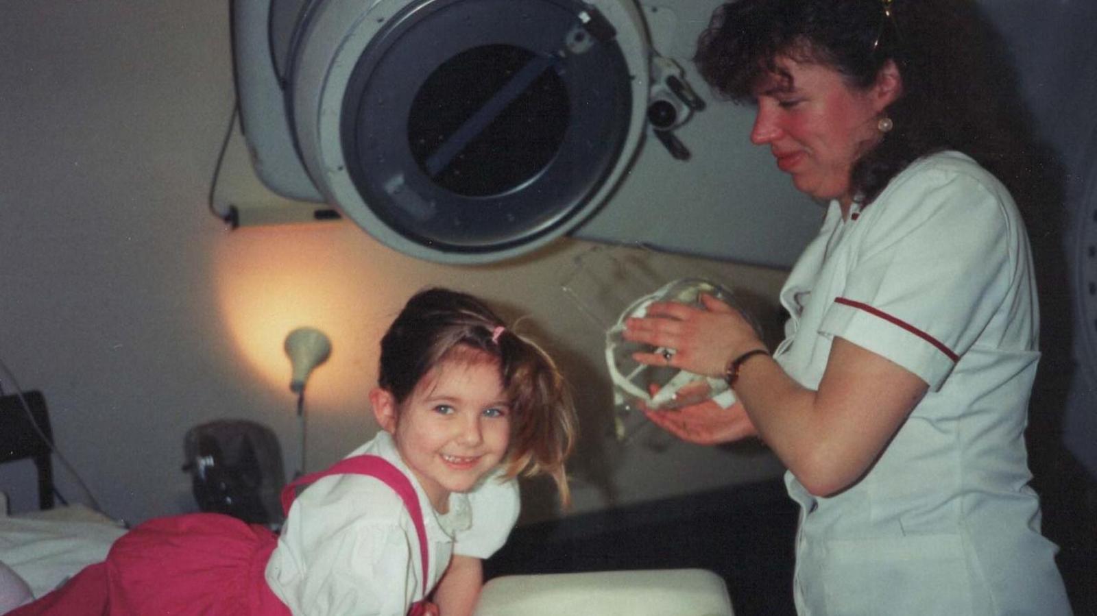 A four-year-old Kat Watson-Wood in hospital wearing her hair in a high pony wearing a white shirt and hot pink skirt smiling. A nurse with long brow curly hair smiles looking at the youngster. 