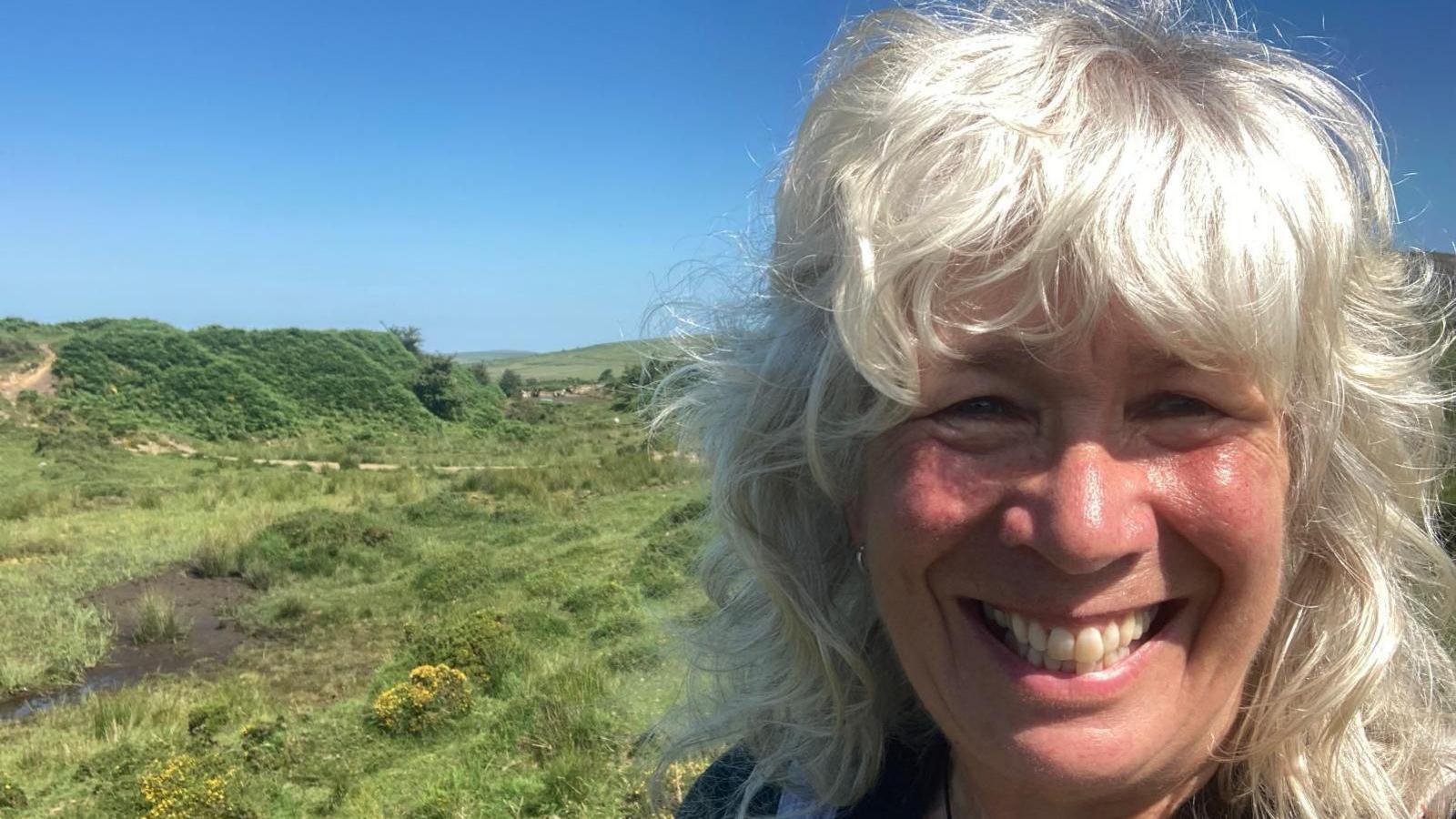 Naomi Wright on the right smiling at the camera. Behind her, on the left, is peatland which is green with an area of mud.