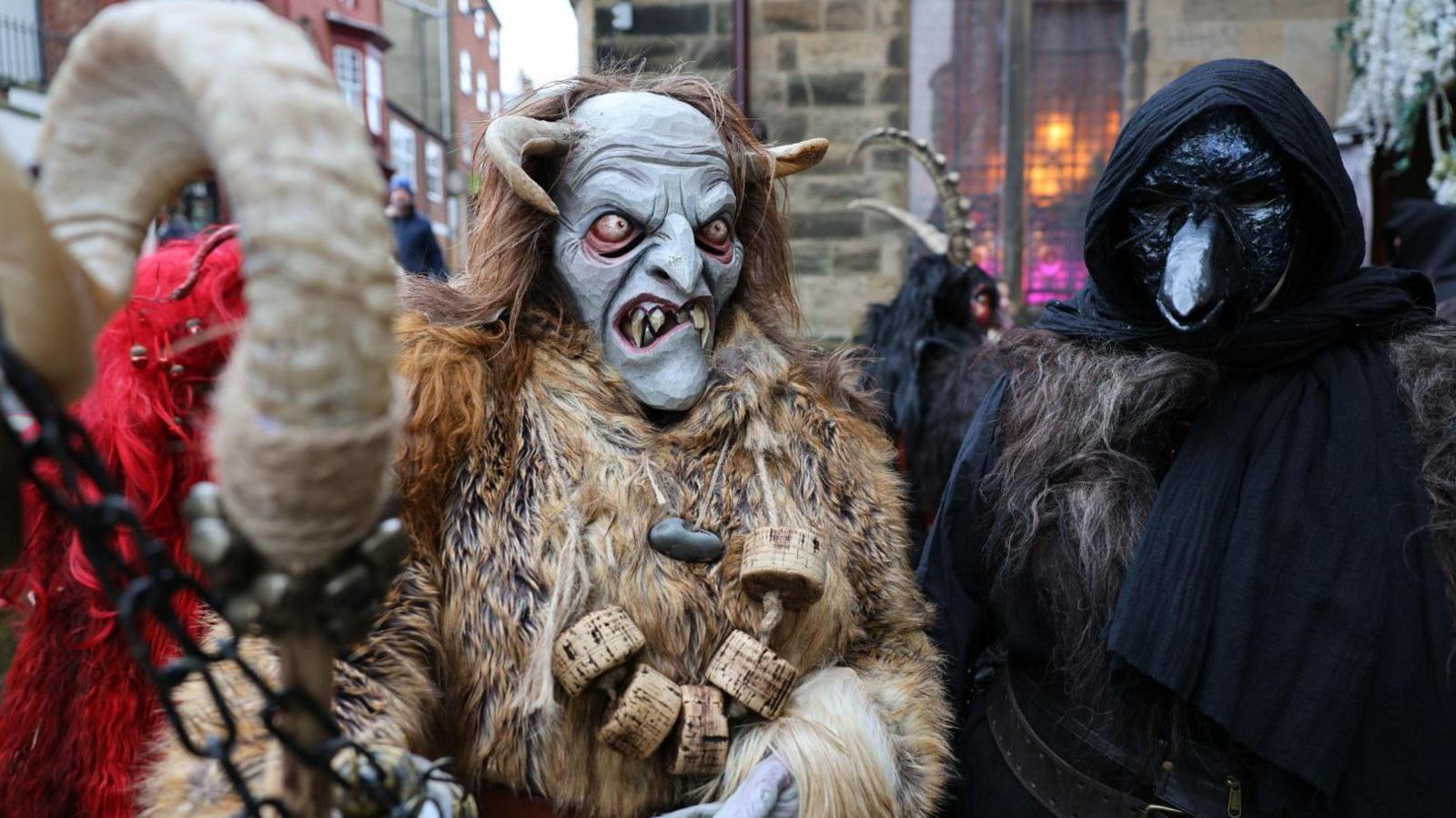 Two people in masks perform as part of the Krampus festival. One is in black with a long beak mask and another is in fur with a white mask and horns.