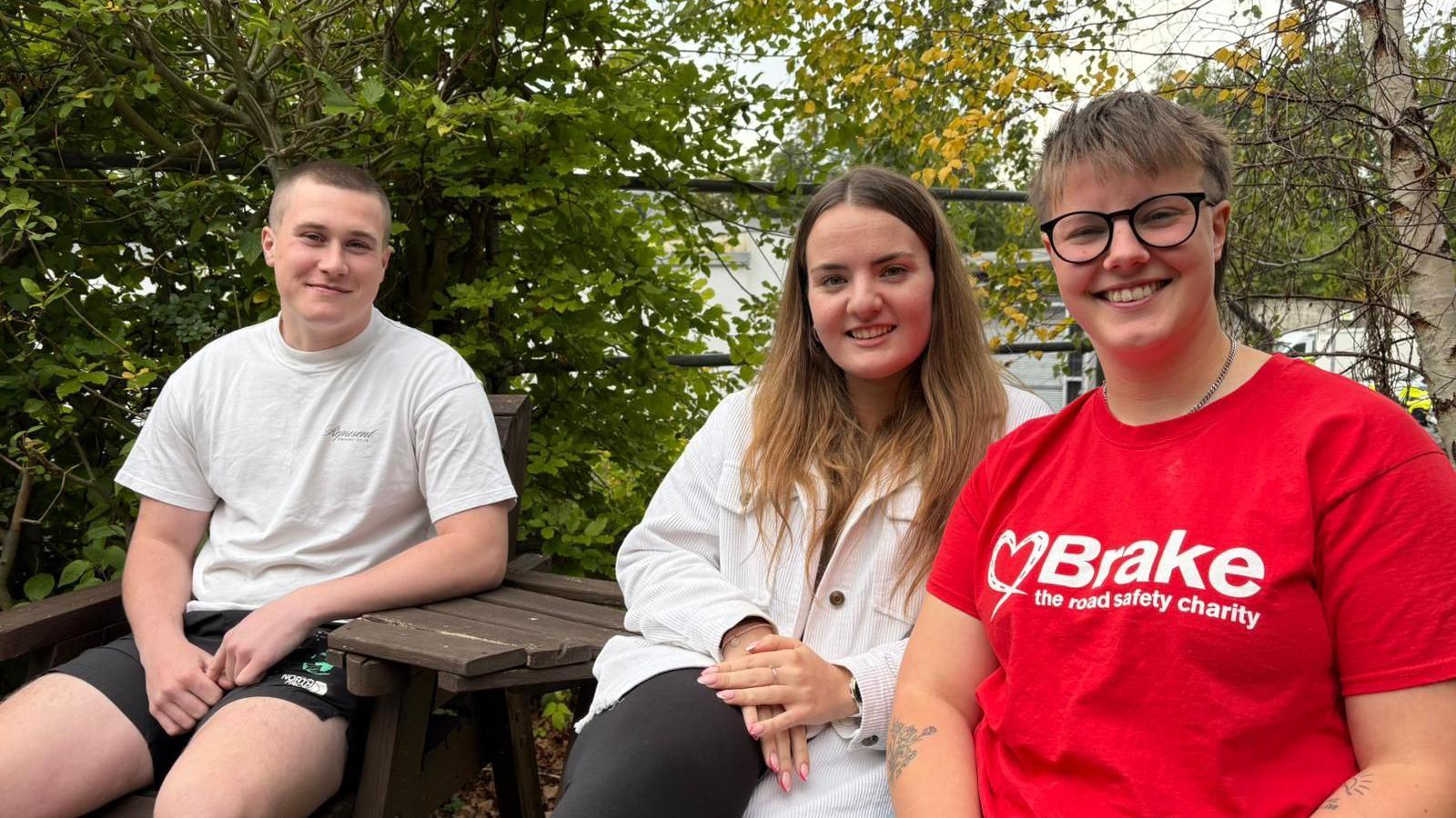 Three friends sit at a picnic table, surrounded by trees. The mood is casual and relaxed. The friend nearest the camera wears a t-shirt with the logo of Brake, the road safety charity.