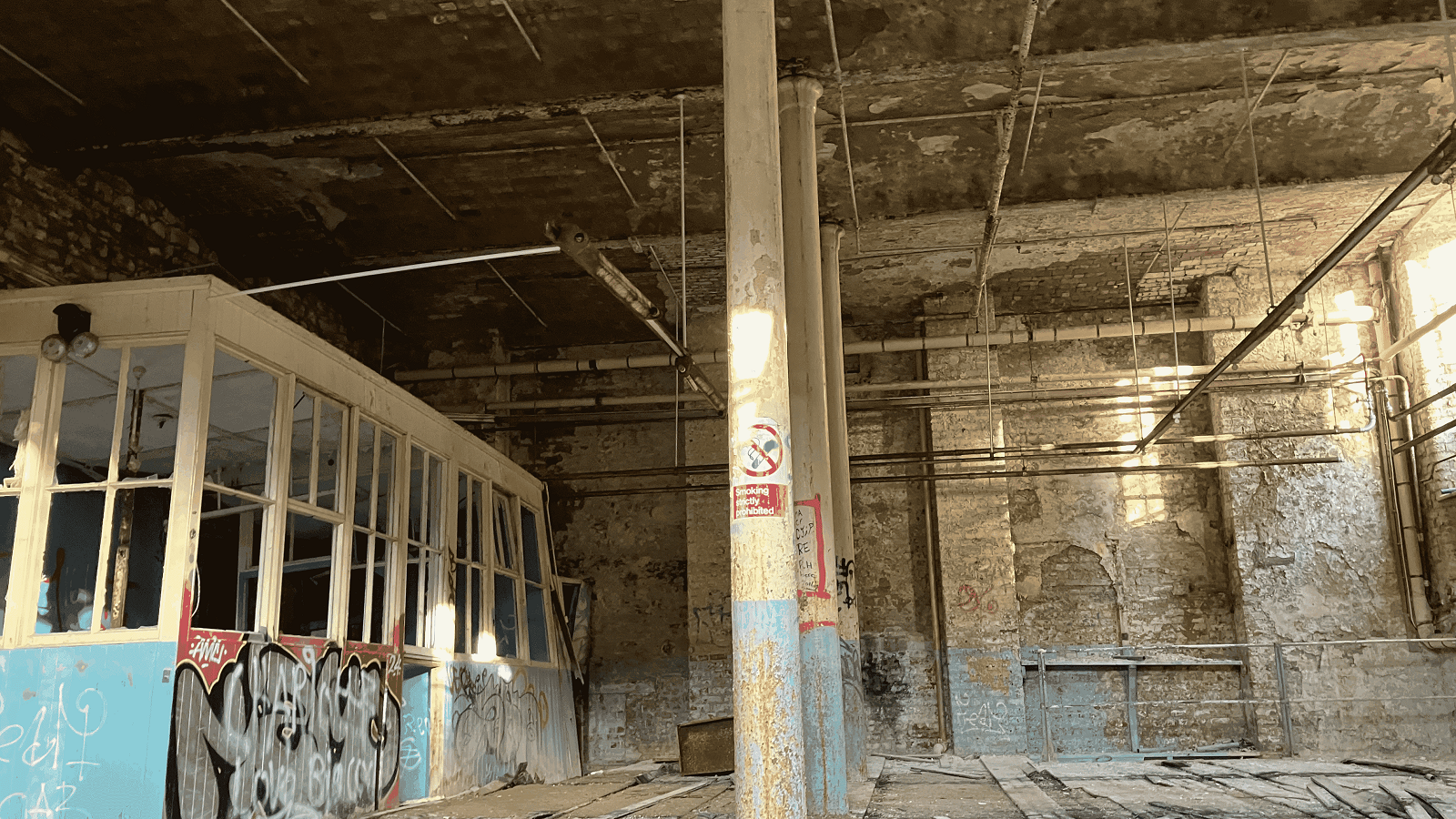 An image of inside one of the many buildings that makes up the Hilden Mill site. Pillars can be seen in the middle of the room, with what used to be an office on the left hand side. The walls are covered in blue paint and graffiti. Exposed pipes run along the ceiling and exposed floorboards decorate the floor.