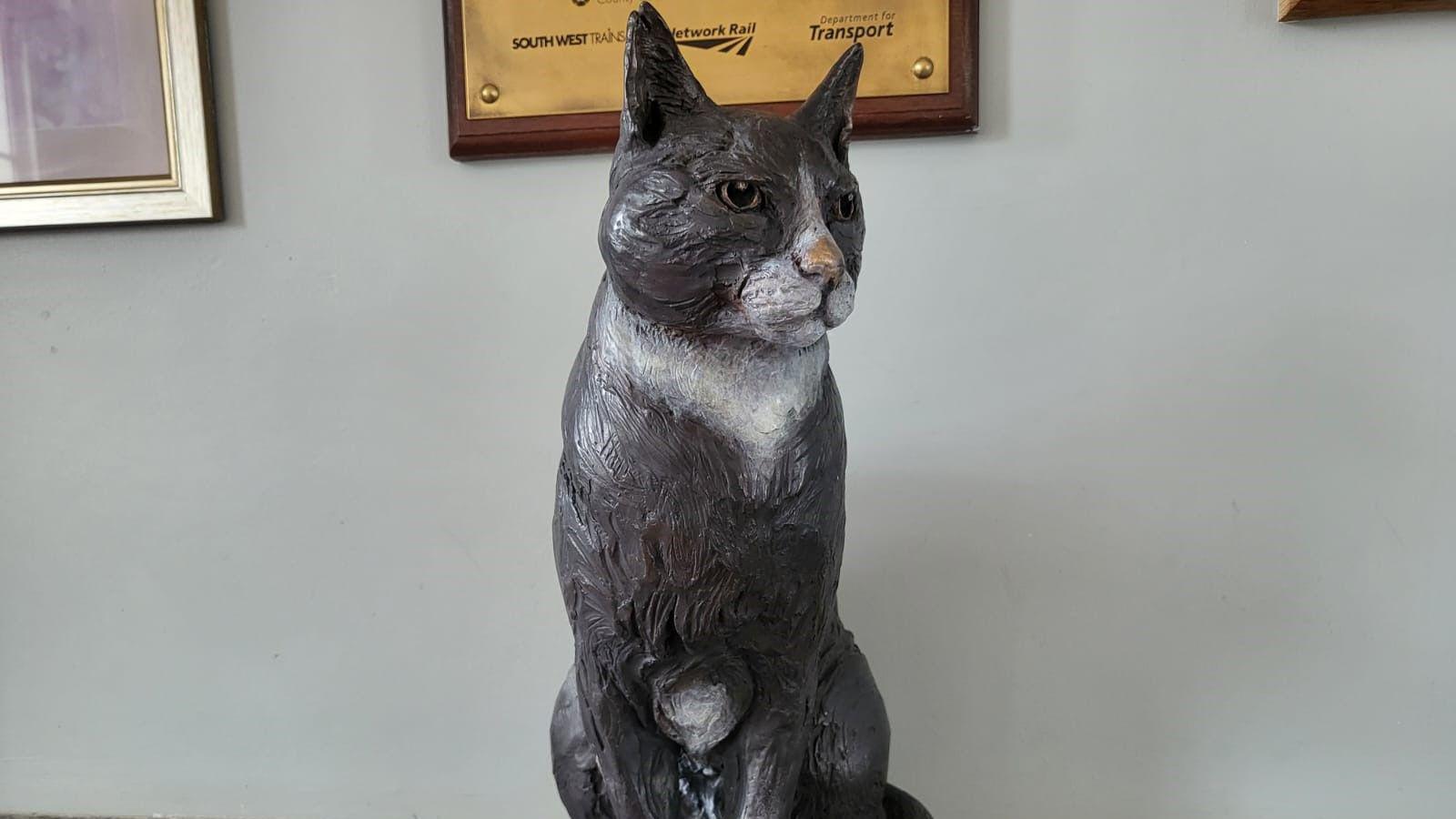 A close-up shot of a life-size statue of a black and white cat against a grey wall, with the bottoms of plaques and pictures just visible above it