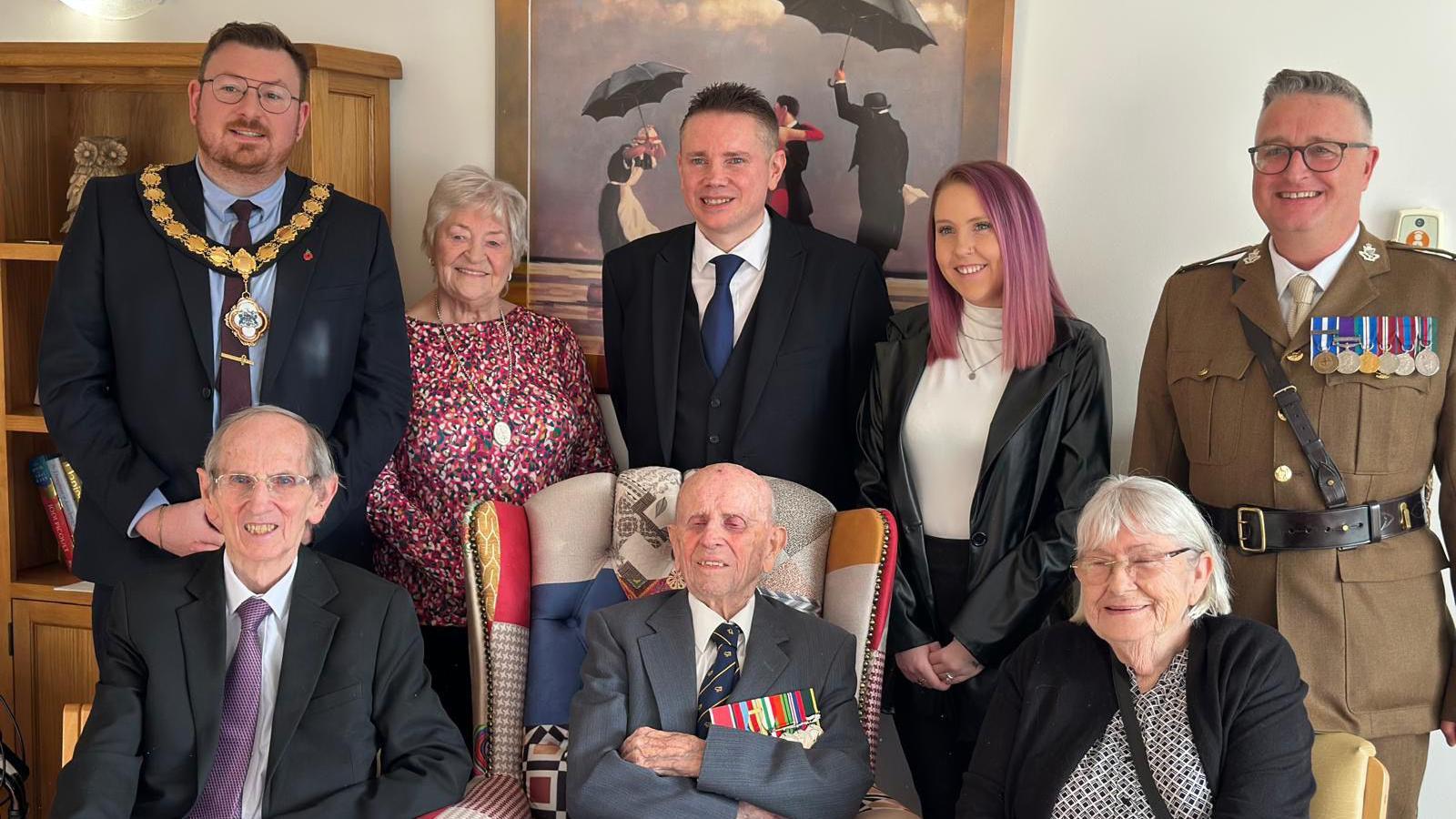 Mr Irwin sits amongst family, friends and military representatives. From left to right, standing up: A man with short dark hair, glasses and beard wearing a navy suit, blue shirt and dark tie. A lady with short grey hair and a flowery blouse. Mr Irwin's grandson with short dark hair black suit, white shirt and blue tie. A lady with purple hair, white turtleneck and black leather jacket. A man with short grey hair and black glasses wearing a dark brown military uniform with his medals. Sitting down, from left to right: an elderly man with short grey hair, metal glasses, wearing a black blazer, white shirt and purple tie. Mr Irwin who is bald and wearing a white shirt, patterned tie and grey blazer with his medals. Then a lady with shoulder length grey hair, silver glasses, wearing a black cardigan and patterned blouse.
