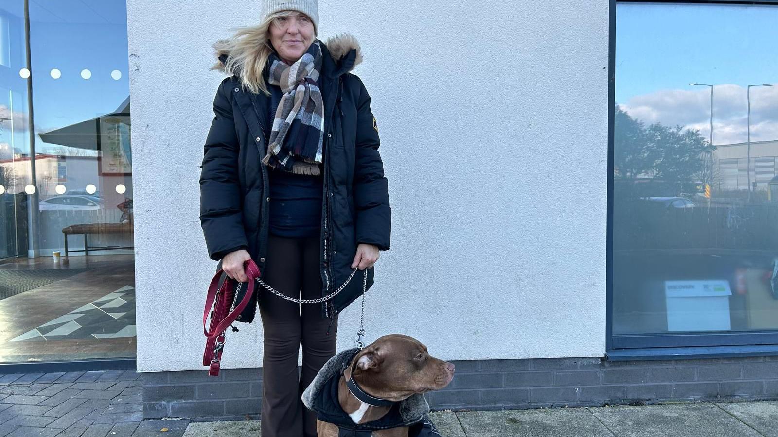 A woman in a light grey woollen hat, chequered scarf and black cold stands by a wall holding a brown bulldog on a lead. 