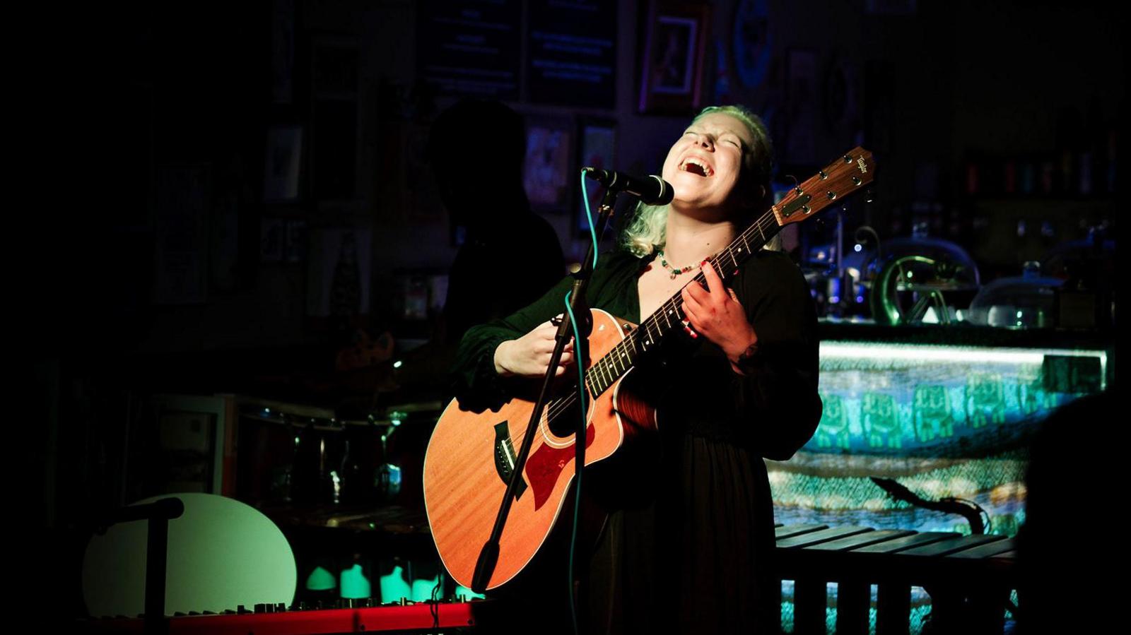 Athena Aperta sings on stage with guitar in hand. The room is dark behind her and her face and the guitar are lit with a spotlight. 