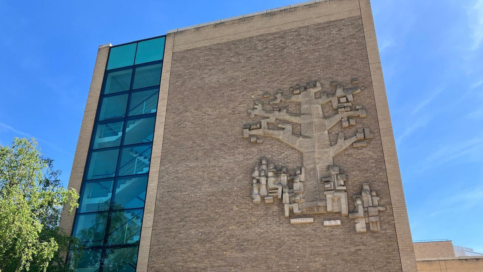 View from the road looking up at the building on a sunny day. It is tall with light bricks and glass from the ground floor up, on the left side of the building, to show a staircase. On the right side is the large sandstone sculpture about the height of four of the short staircases . It is blocky and shows people sitting under an oak tree. 