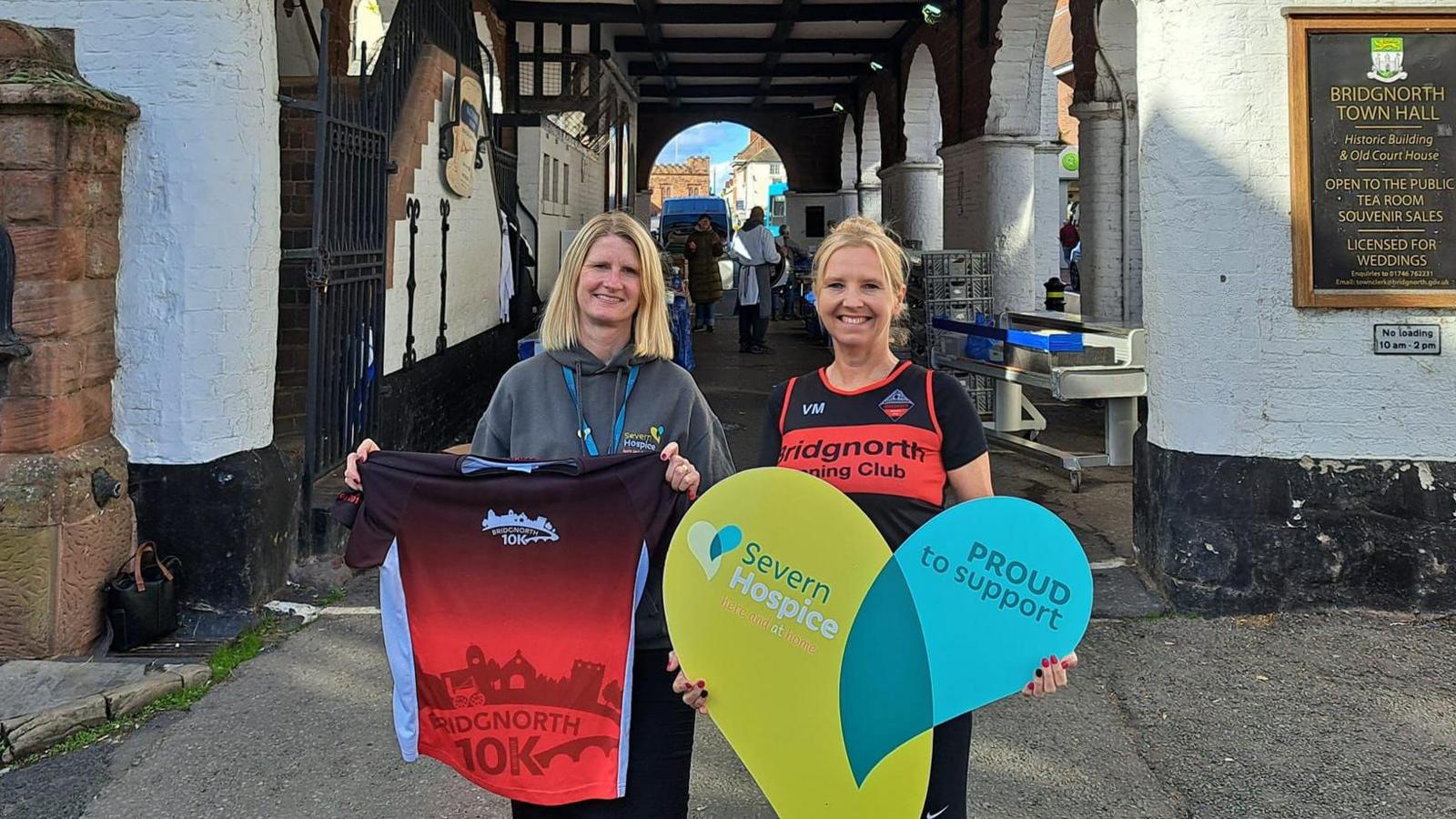 Two women stand side by side under an archway. The woman on the left is blonde and wearing a grey hoodie. She is holding up a Bridgnorth 10k t-shirt, which is predominantly red. The other woman is also blonde and wearing a black and red Bridgnorth running club vest. She is holding a heart-shaped sign saying Proud to Support Severn Hospice.