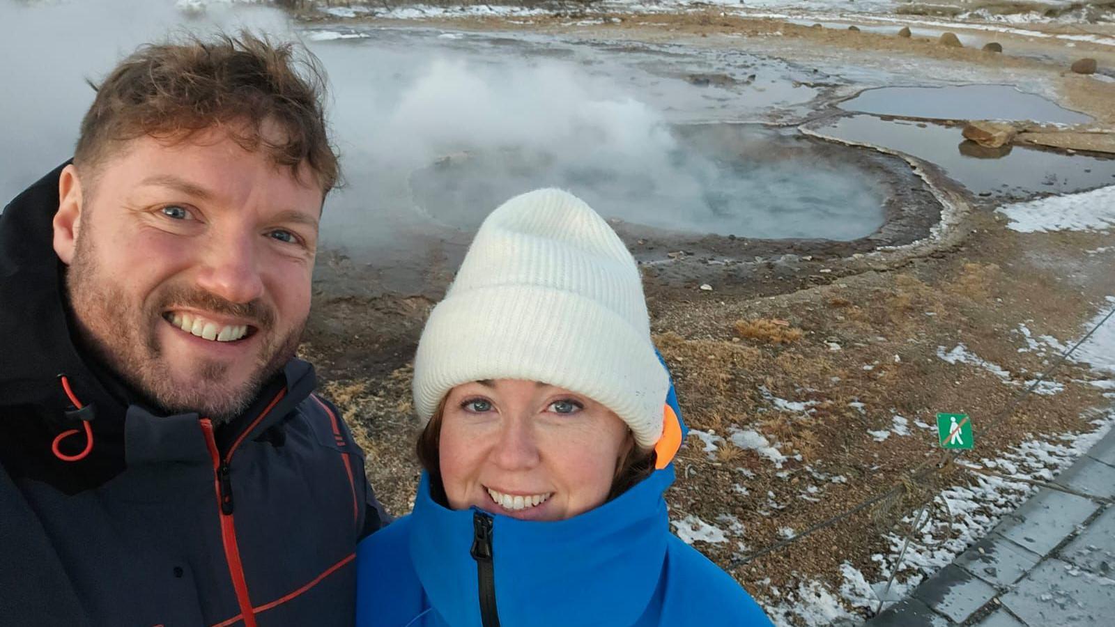 A man and a woman wearing warm winter clothing stand in front of steam rising from the ground in Iceland