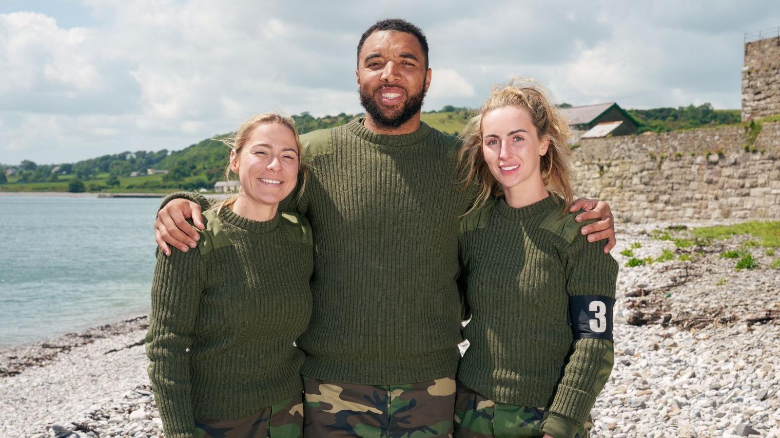 Lucy Spraggan, Troy Deeney and Michaella McCollum stand on a beach with their arms around each other, smiling at the camera. The three are wearing green army style jumpers and camo trousers.