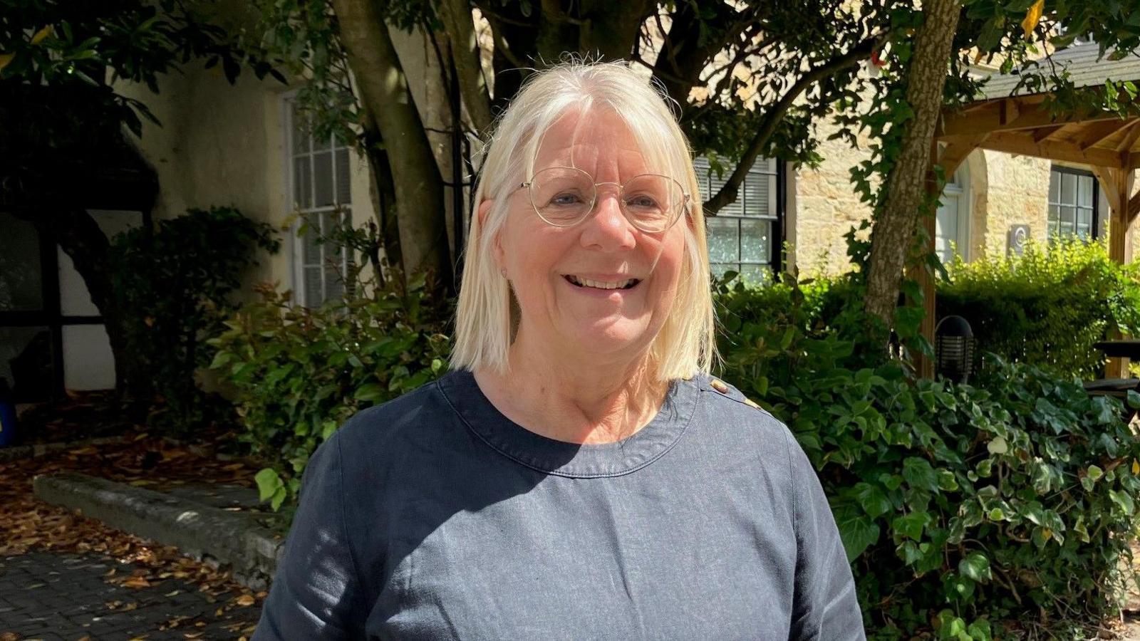 A head-and-shoulders image of a smiling Ruth Huxley. She is wearing a navy blue top and has blonde hair in a long bob and glasses. She is standing in front of a cream-coloured building which has trees and a bush in front of it.