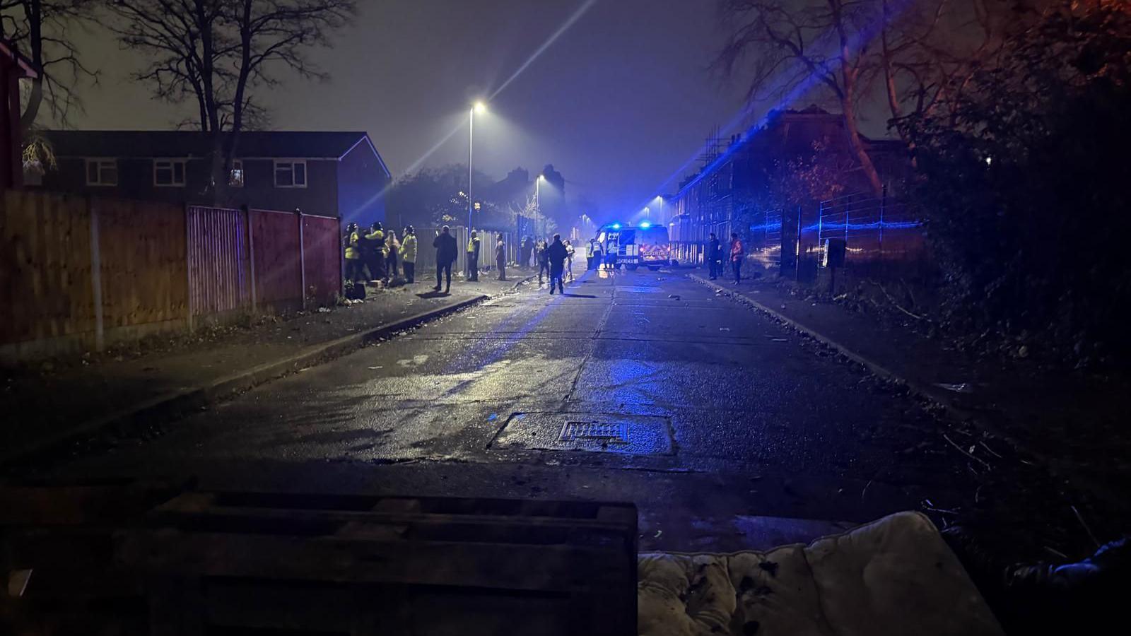 A police van and police officers wearing hi-vis jackets at the scene of the attack, with a number of other people standing in the street.