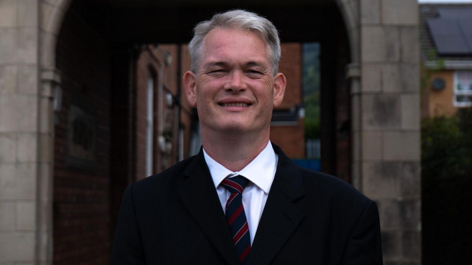 Richard Tunnicliffe, wearing a black jacket, dark blue and red tie, and white shirt, in front of a stone arch.