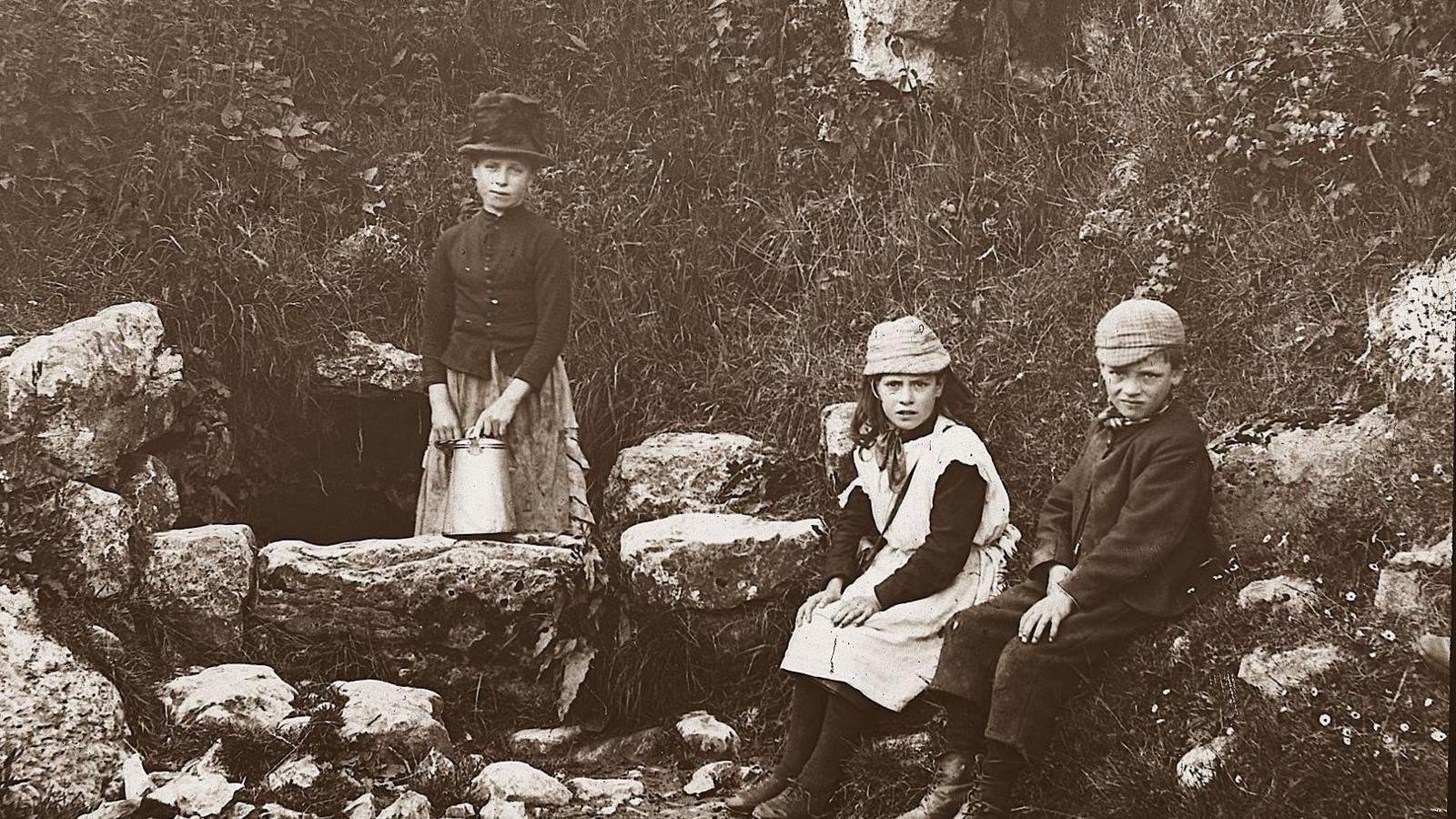 Three Victorian children sit beside a stone well. It is a historic, black and white image.