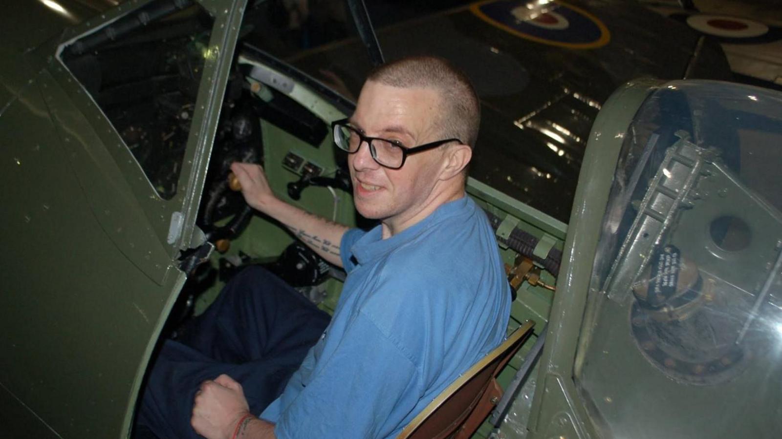 Michael Webb has square black glasses and close shaved hair. He is wearing a blue top and is sitting in the cockpit of an old RAF aircraft.