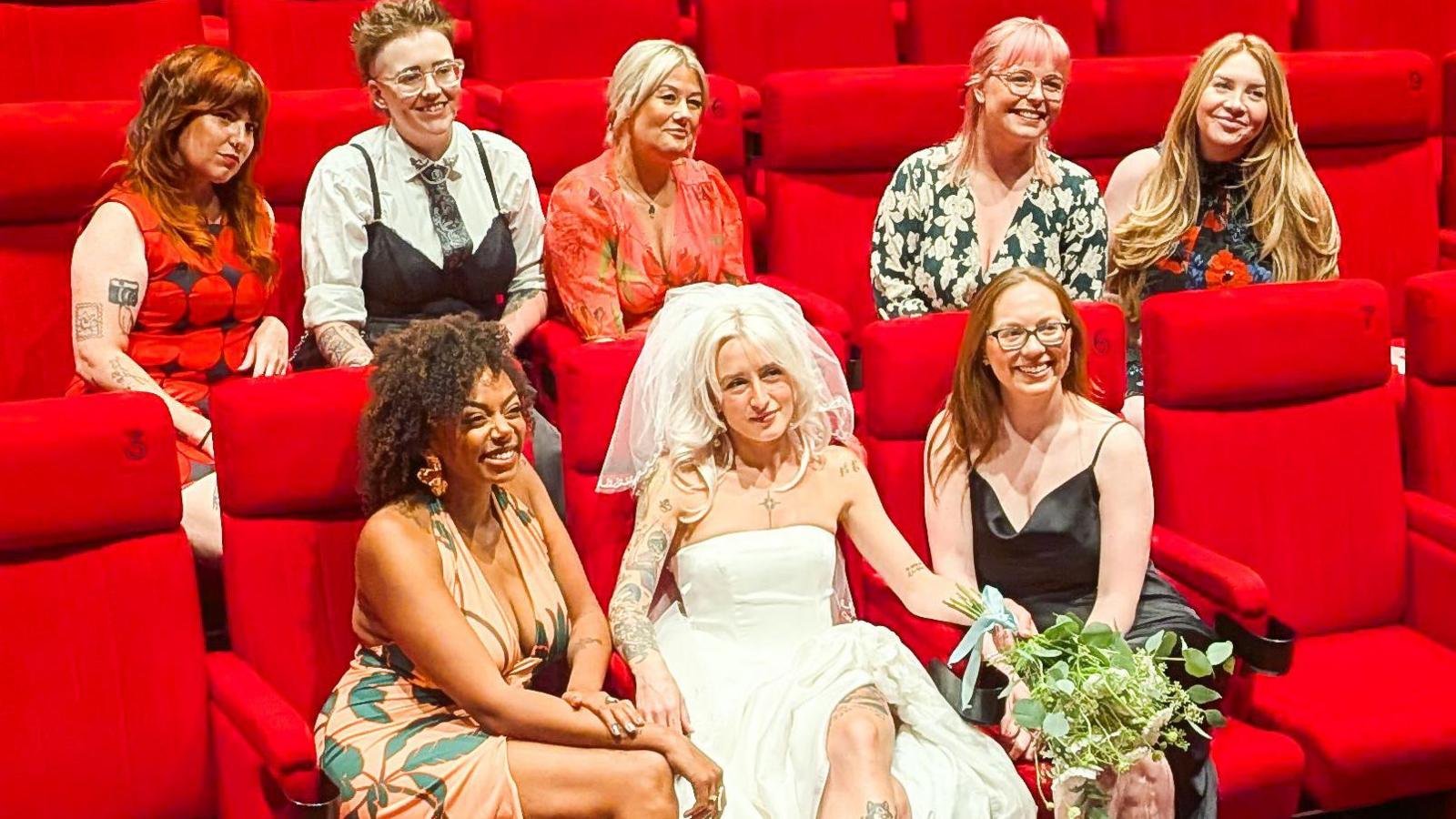 A group of women wearing wedding formal wear are smiling while sitting on red cushioned seats. The bride is wearing a strapless white wedding dress and a veil and is holding a bouquet.