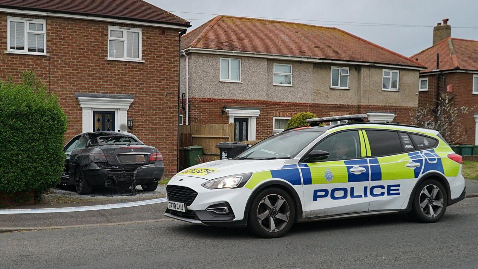 A police car is pictured in front of some police tape and a burnt out black Mercedes on a residential street in Eastbourne