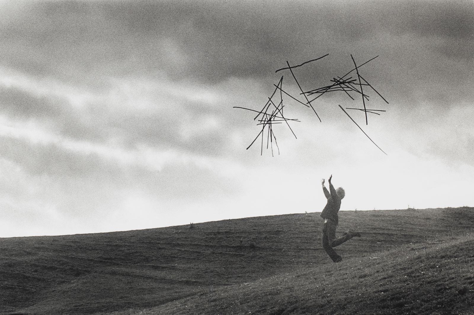 A man caught in action throwing sticks in the air. The backdrop is a cloudy sky with the man standing on a hill. The image is black and white.