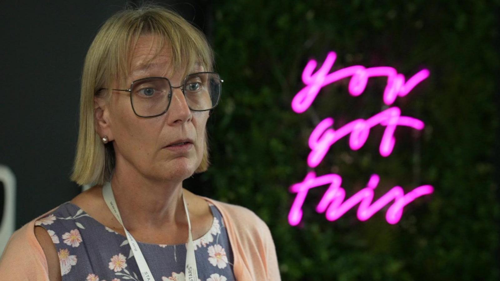 Helen Stevens has shoulder length blonde hair and glasses. She is wearing a blue top with white flowers and a peach coloured cardigan. She has a white lanyard around her neck. She is sitting in front of a green wall with a neon pink sign that says "you got this".