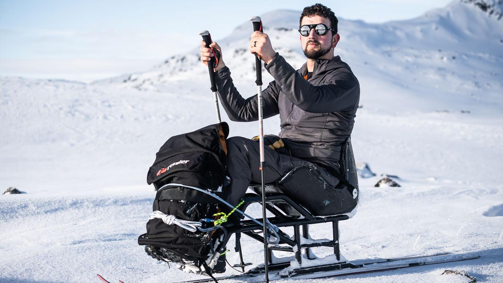 Aman sits on a sled with skis attached to the bottom while he holds two ski poles. He is in the middle of a snowfield with snowy hills in the background.
