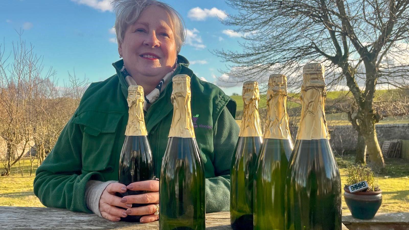 Lorna Jackson, who has grey hair and is wearing a green jacket with a scarf holds on to one of five bottles of sparkling wine. There are trees and fields in the background