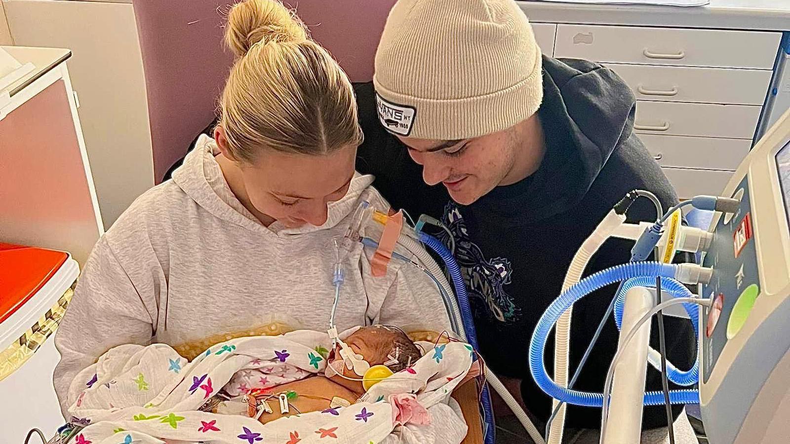 Megan is wearing a grey hoody and has her blond hair pulled back in a messy bus. She is cradling a small newborn in her arms who is lying in a white blanket or muslin which has colourful stars on it. The baby has various medical wires attached to her and in the foreground part of a medical device on a stand can be seen. Megan is sat next to her partner who is wearing a black hoody and cream beanie. Both parents are looking down at the baby