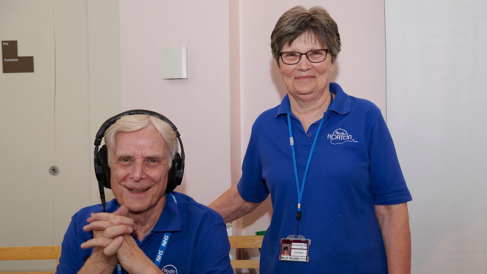 Tony Wilson who has short white hair and is wearing a blue t shirt and headphones, sat down next to a woman stood up wearing a blue t shirt, with glasses and grey hair