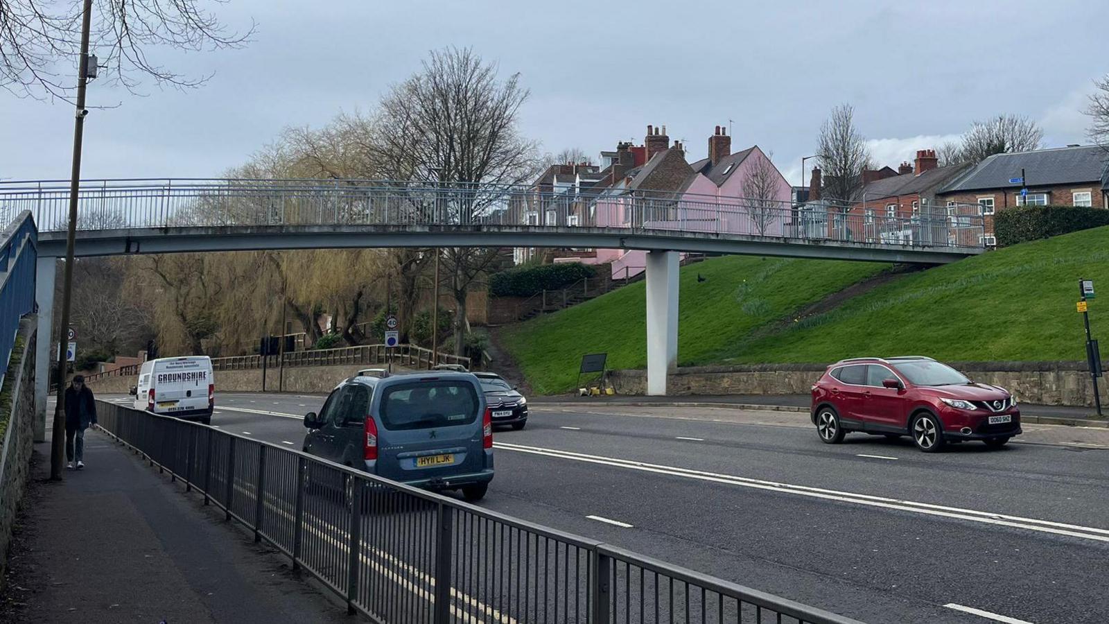 The footbridge has metal railings and a dual carriageway road beneath it.