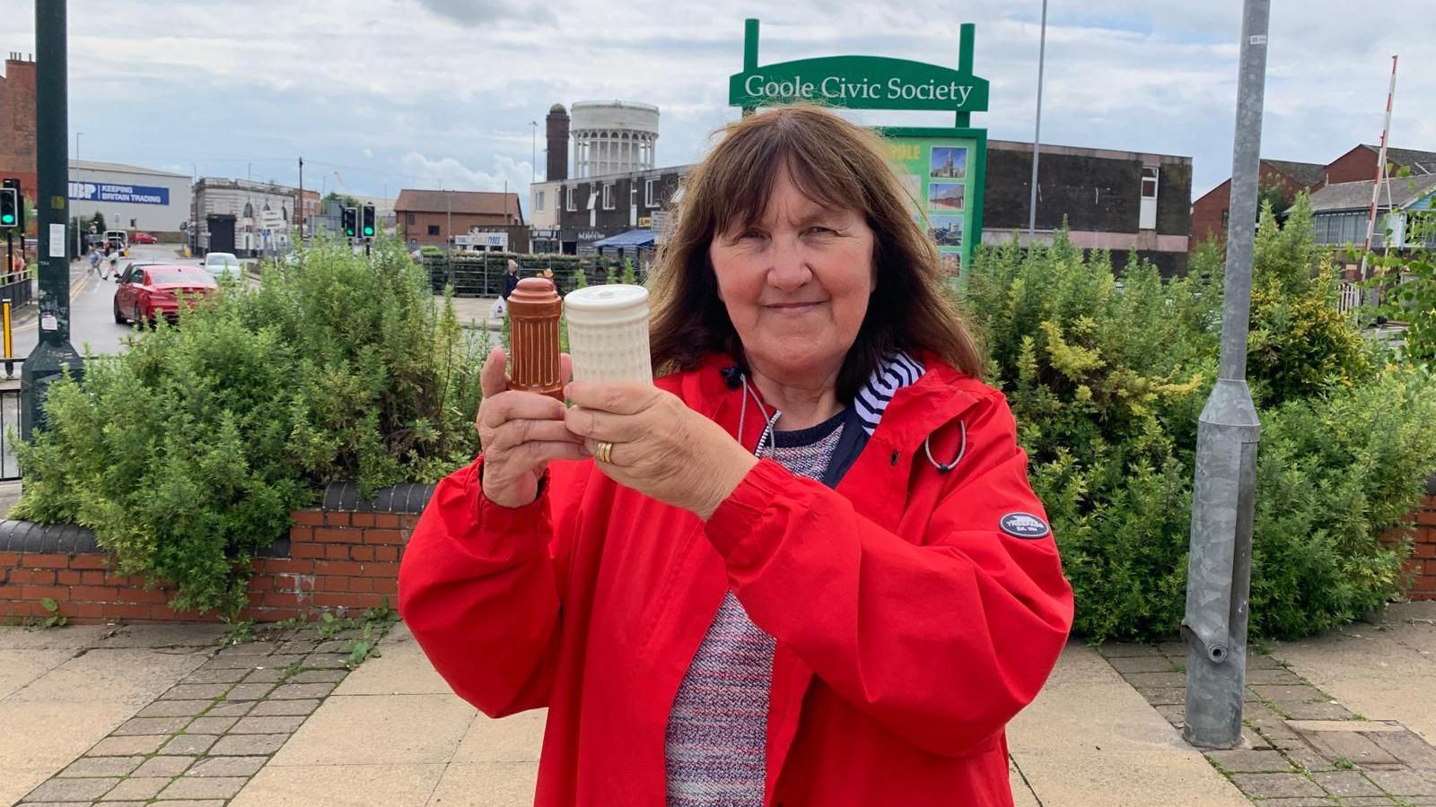 Margaret Hicks-Clarke wearing a red coat and with brown shoulder-length hair holds up the ceramic salt and pepper pots. They resemble the town's twin water towers which are behind her in the picture. 