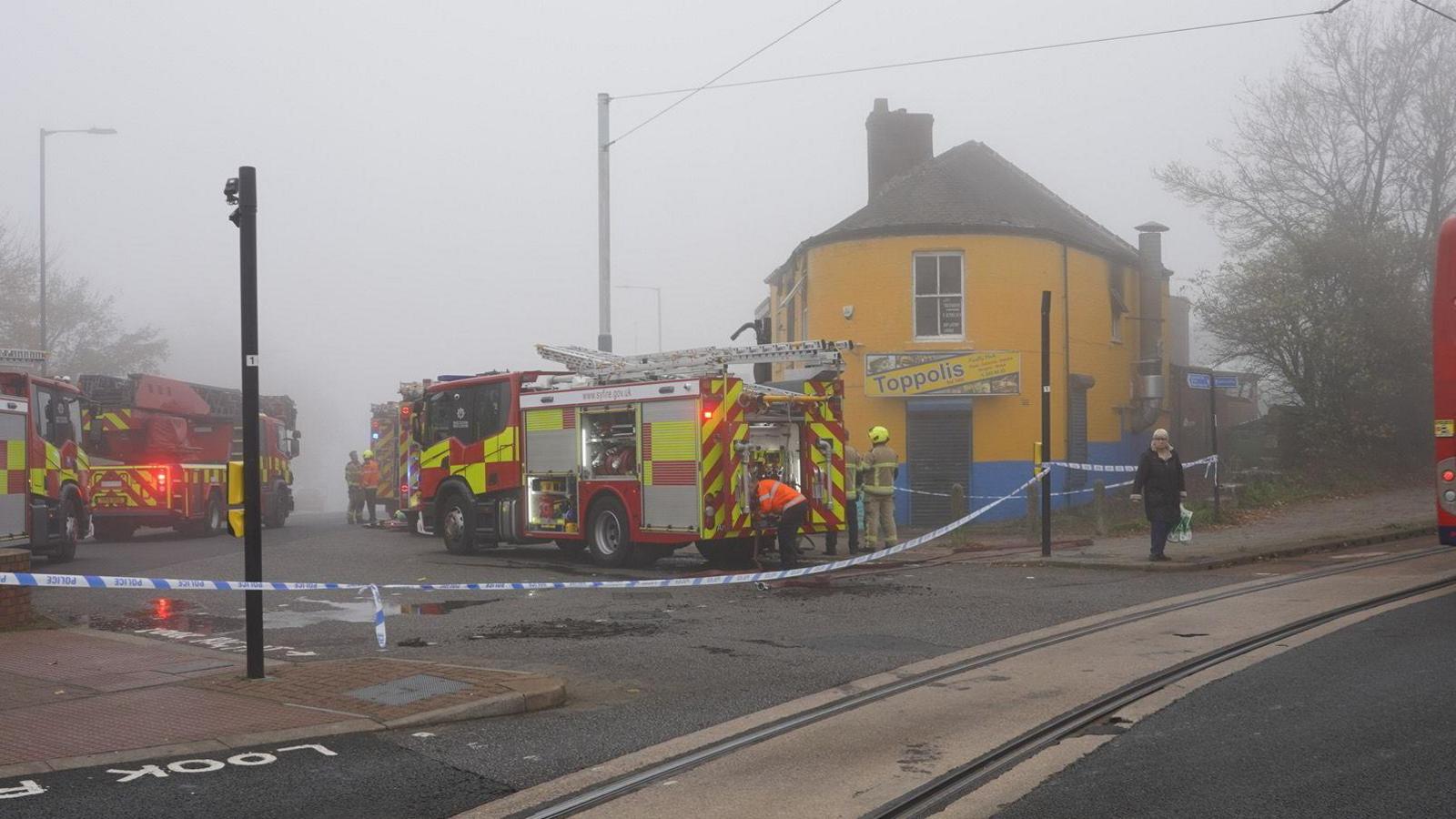 Fire engines next to a yellow building. There is blue police tape stretched across the junction of the road