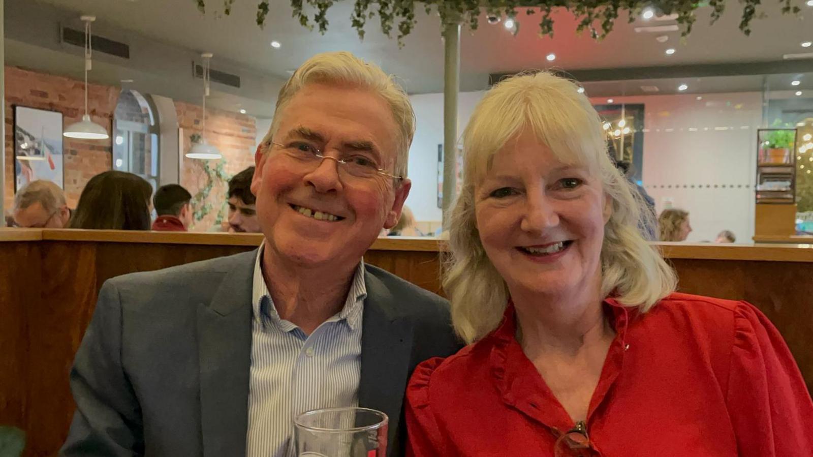 A photograph of a couple in a restaurant. The man on the left wears a shirt and a suit and the woman is wearing a red blouse. 