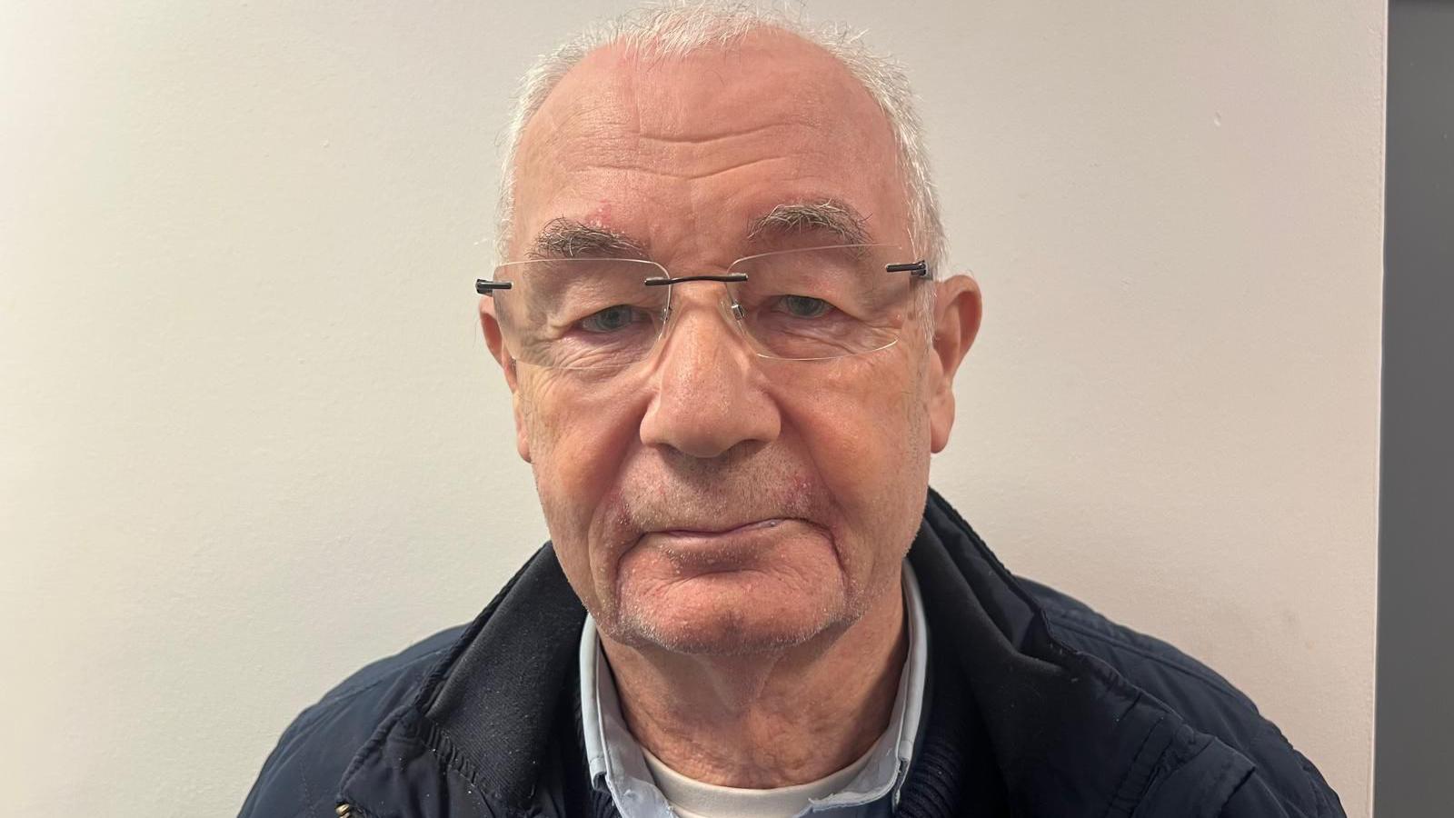 Mickey McKinney is wearing a blue shirt, white top and a navy coat. He has glasses on and short grey hair. He is in front of a white wall.