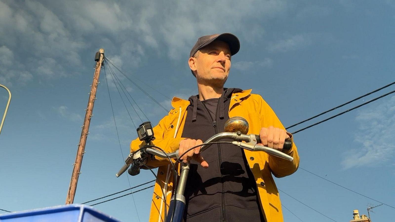 Paul Troop is sat on his bike with his hands on the bars. He is looking in the distance wearing a yellow coat.