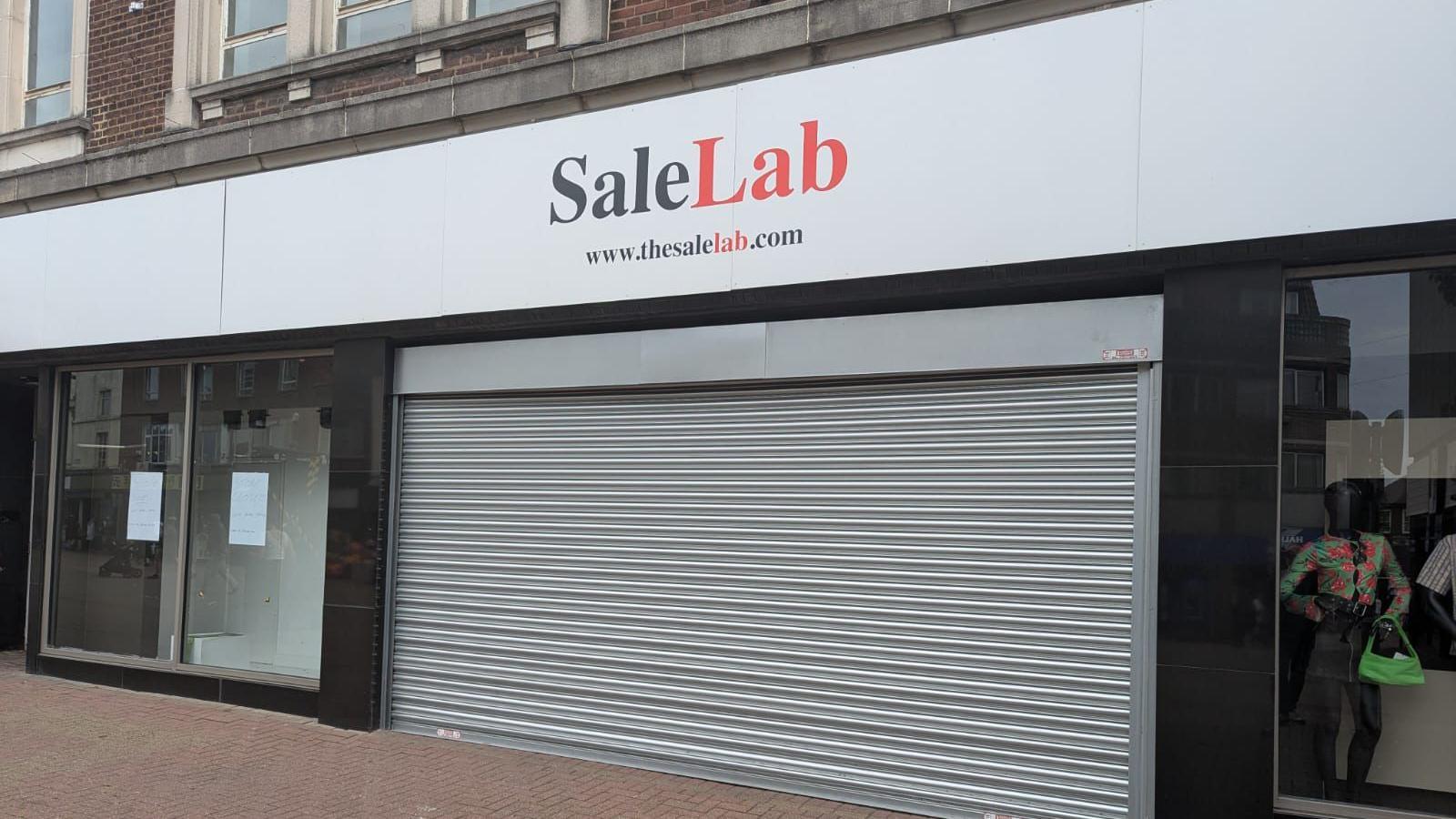 A shop front with shutters down. There is white sign with red and red lettering that read Sale Lab.