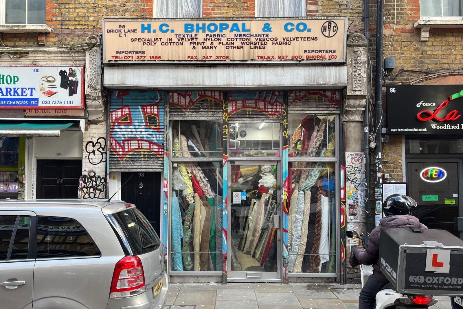 A parked car and a delivery driver on a moped parked outside a shop with numerous rolls of textiles in the windows