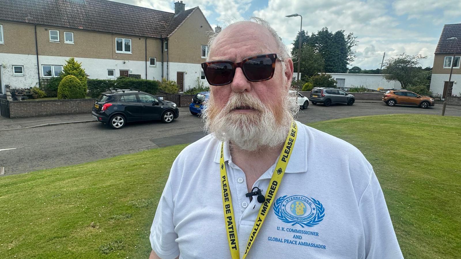 Bill Mackie, 71, with short white hair and a white bears, wears dark glasses for his eye condition and a military polo shirt. He wears a lanyard saying "please be patient, visually impaired". The houses of the estate lie behind him on two sides.