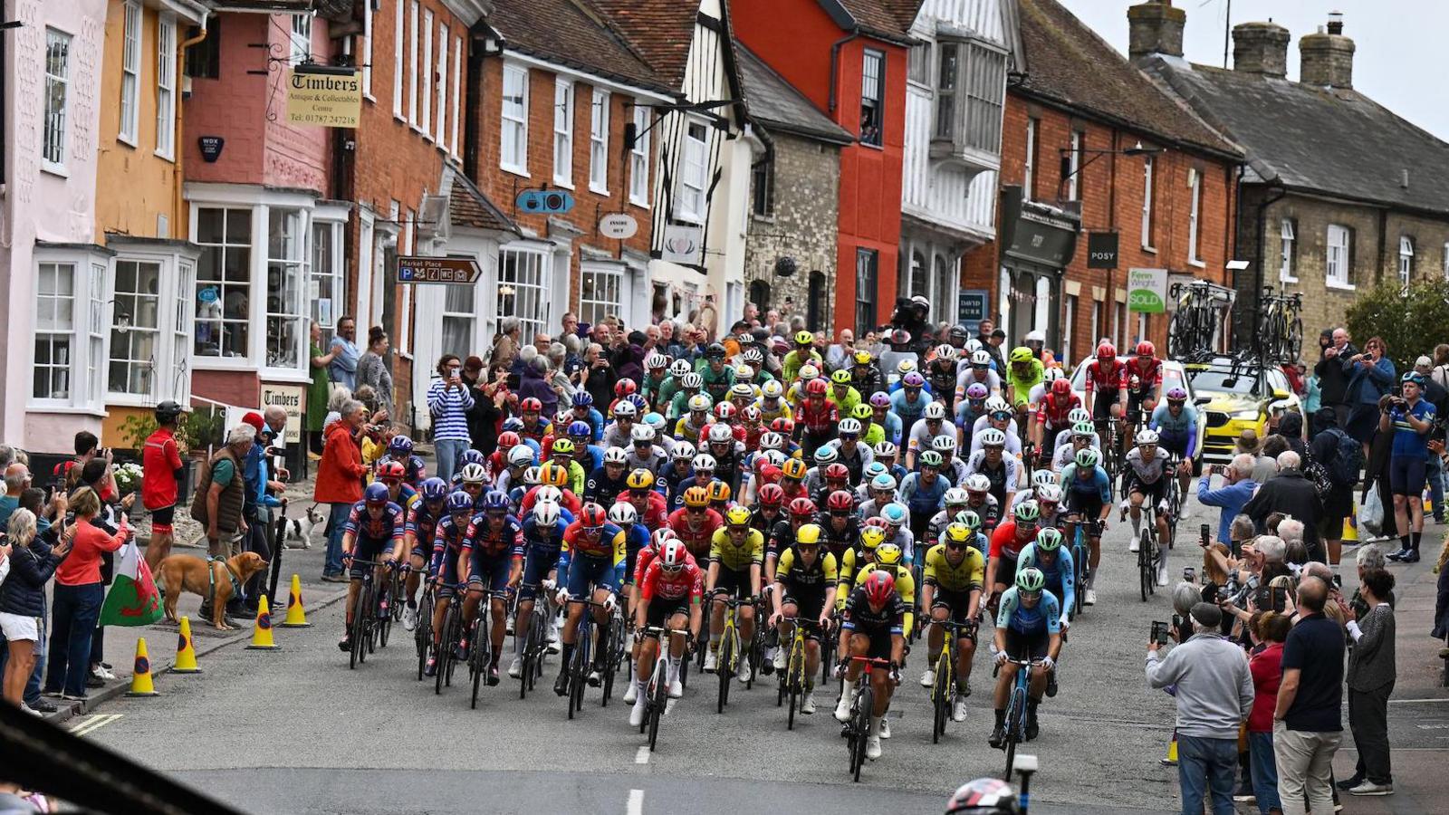 A large group of cyclists all wearing different coloured jerseys ride down a hill. Spectators watch and cheer them on from the side of the road.