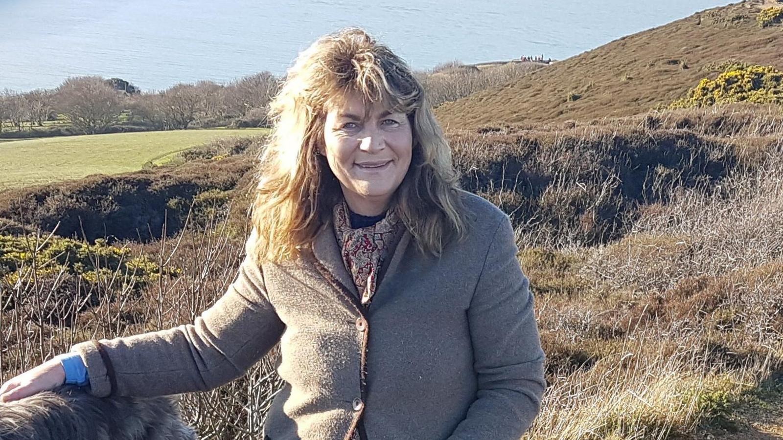 Annabel Harrison stands on a hill with a view of the sea in the background. She has wavy, fair hair and wears a brown jacket and patterned scarf.