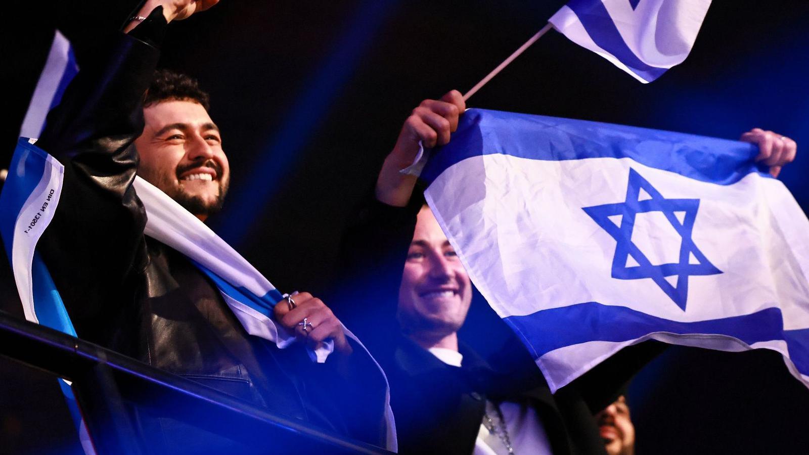 Two people, perched overlooking a metal railing, are smiling and celebrating. The man on the left has short black hair and a black goatee. He is draped in an Israeli flag, which is blue stripes, with a large stripe of white in the middle and a blue Star of David. The woman on the left is holding a small flag of Israel on a stick as well as a larger flag which she is waving in both hands. 