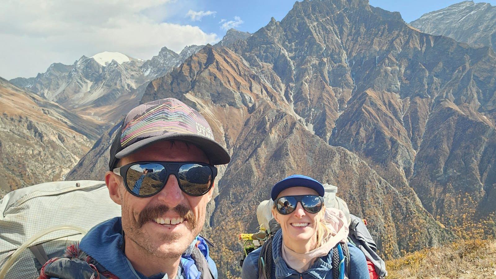 Rik is taking a selfie of the couple and they're both smiling at the camera. They both have large backpacks on with blue fleeces and have sun glasses and caps on. Behind them is a mountain range.