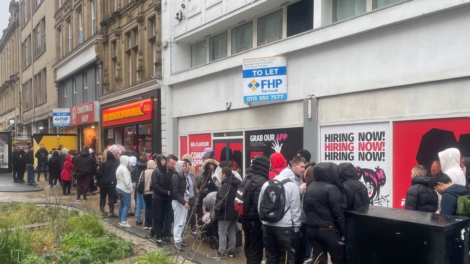 A large queue of people wrapped up in warm clothes stand on a grey day outside a series of shops