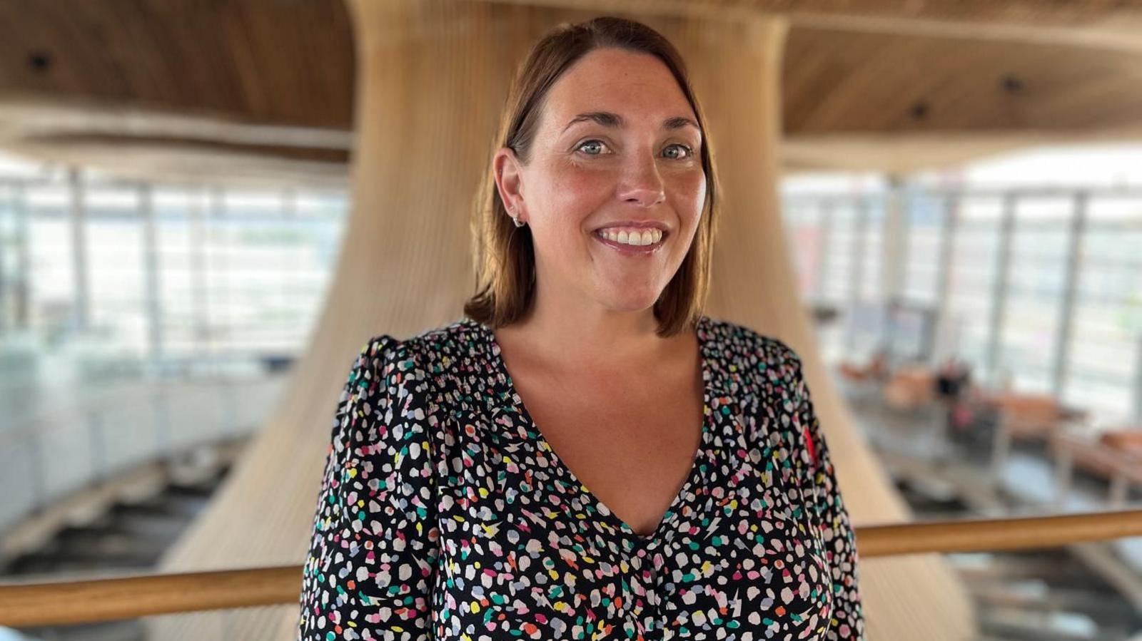 The image shows Heledd Fychan standing in the Senedd looking directly at the camera and smiling. She is wearing a colourful dress. The Senedd's funnel is behind her.
